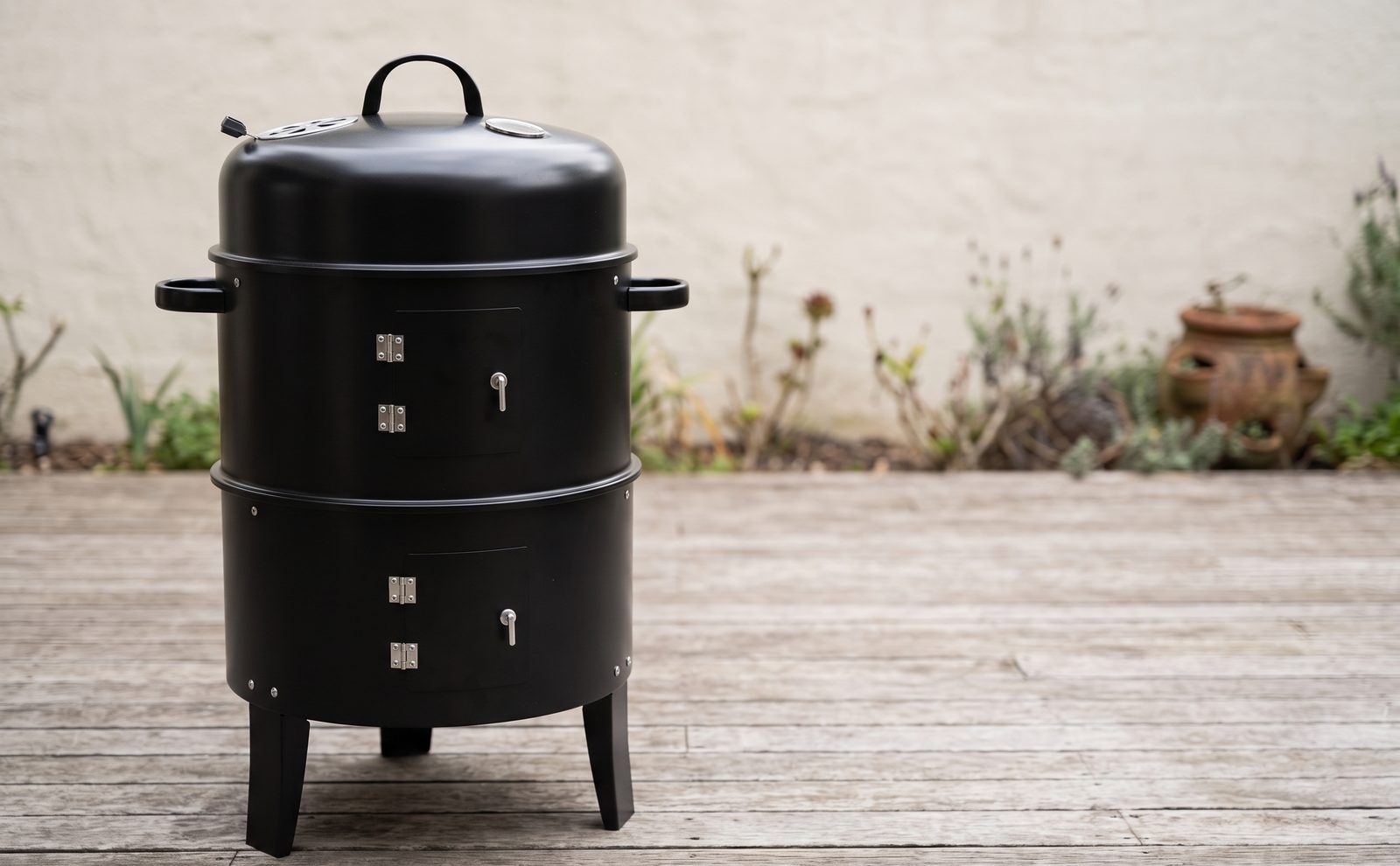 Black charcoal smoker on a timber deck with icons for water pan, grates and lid dial